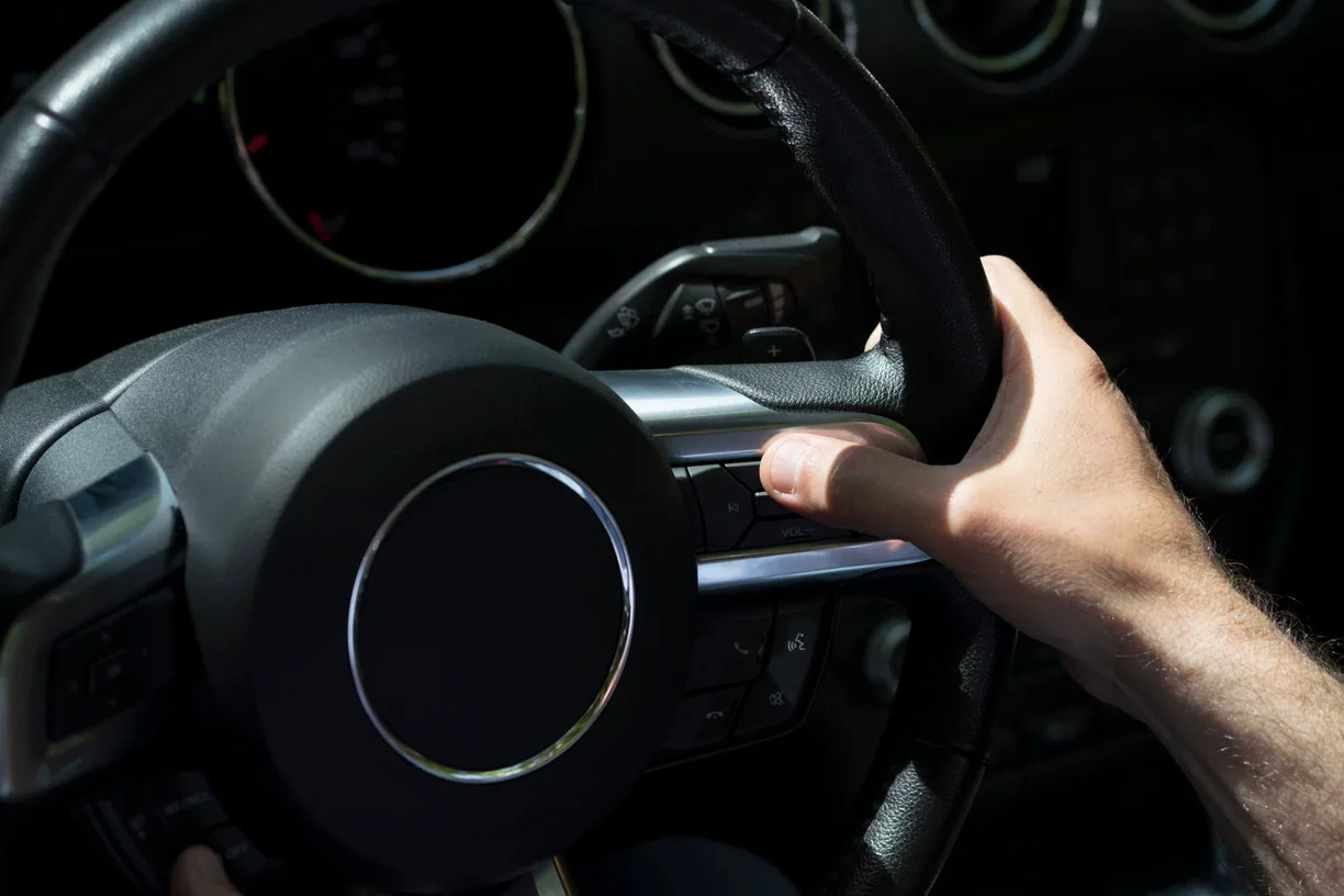 Driver's hand on a steering wheel, instrument cluster lit behind it.