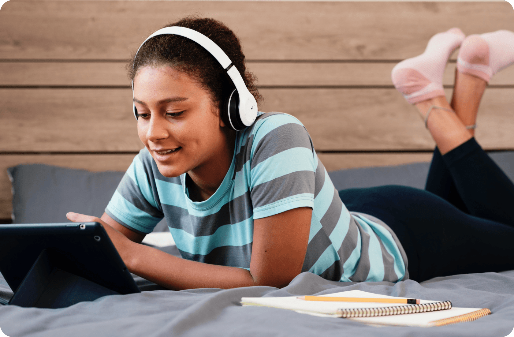 Teenager wearing headphones, lying on bed, using a tablet with notebooks nearby.